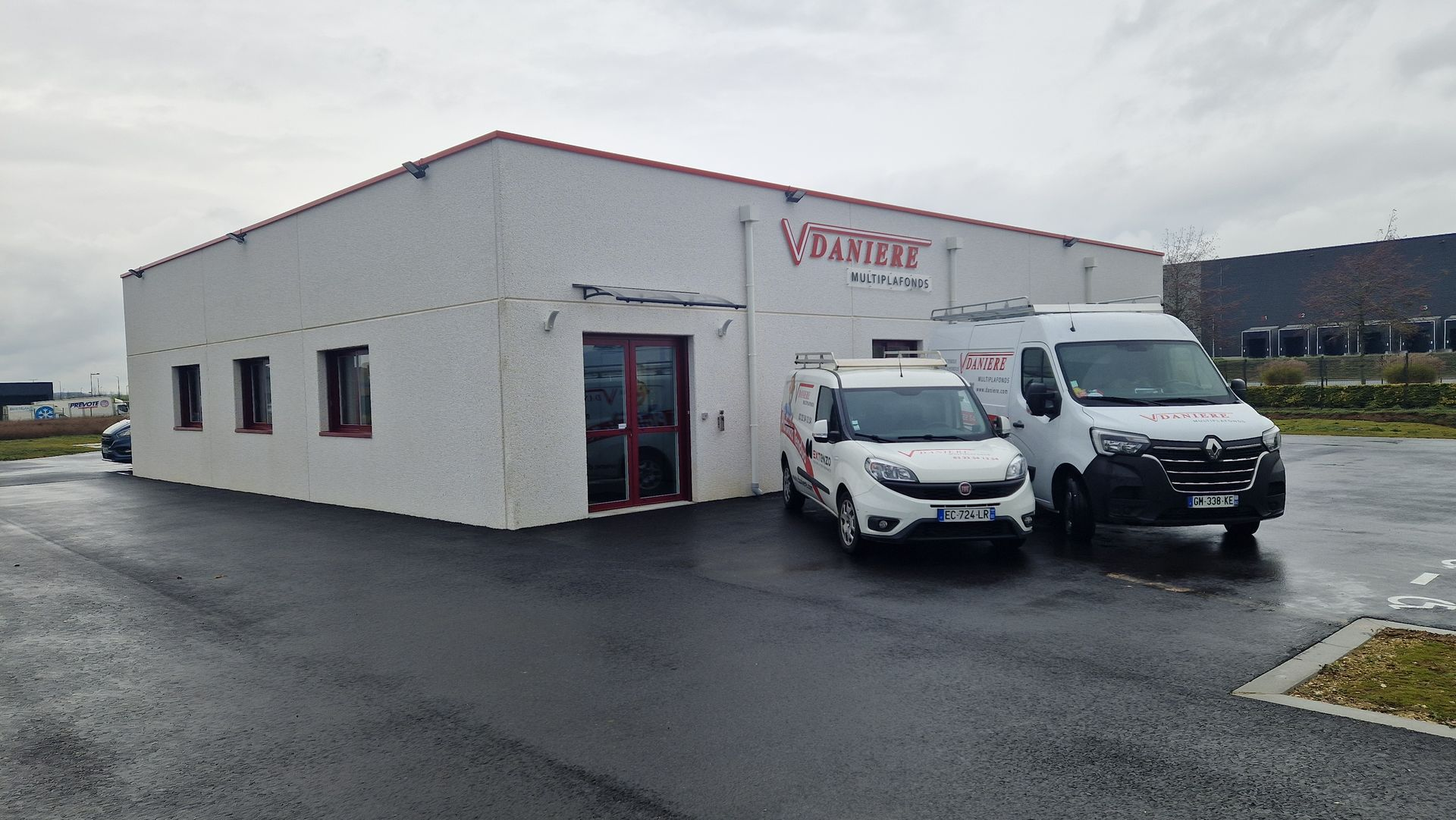 Bâtiment blanc aux moulures rouges. Deux camionnettes garées à l'extérieur sur l'asphalte mouillé.