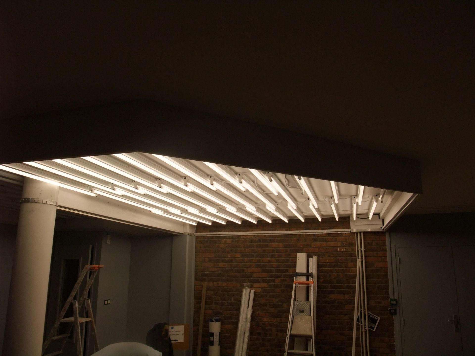 Un plafond avec des lumières horizontales lumineuses est installé dans une pièce inachevée avec des briques et des échelles.