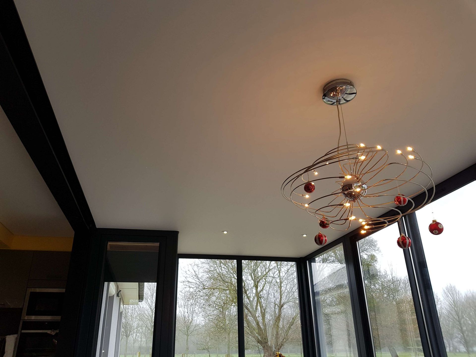Un lustre moderne aux ornements rouges est suspendu à un plafond blanc, près de grandes fenêtres et de poutres noires.