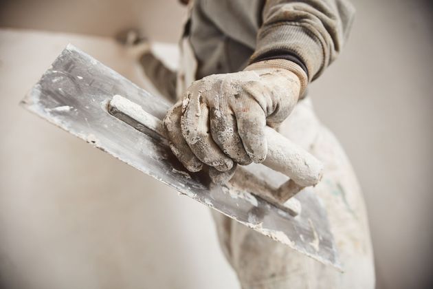 Die behandschuhte Hand eines Arbeiters hält eine mit Gips bedeckte Glättkelle und trägt diesen auf eine Wand auf.