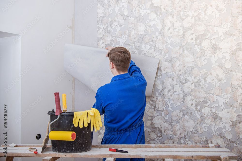 Eine Person in blauem Overall tapeziert eine Wand. Werkzeug steht in einem Eimer daneben.