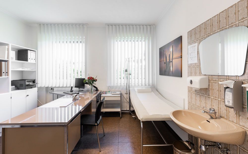 Eine Arztpraxis mit Schreibtisch, Bett, Waschbecken und Spiegel.