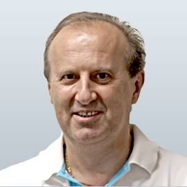 Ein Mann in einem weissen Hemd lächelt und blickt dem Betrachter direkt vor einem hellblauen Hintergrund an.