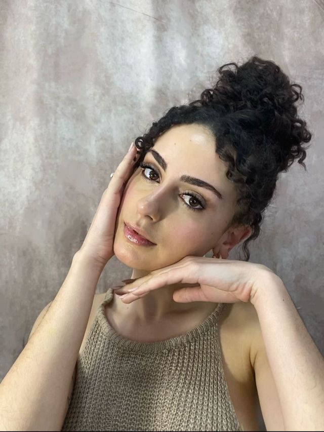 Mujer posando con las manos bajo la barbilla sobre un fondo gris suave, vistiendo una blusa beige y con el pelo rizado recogido en un moño.