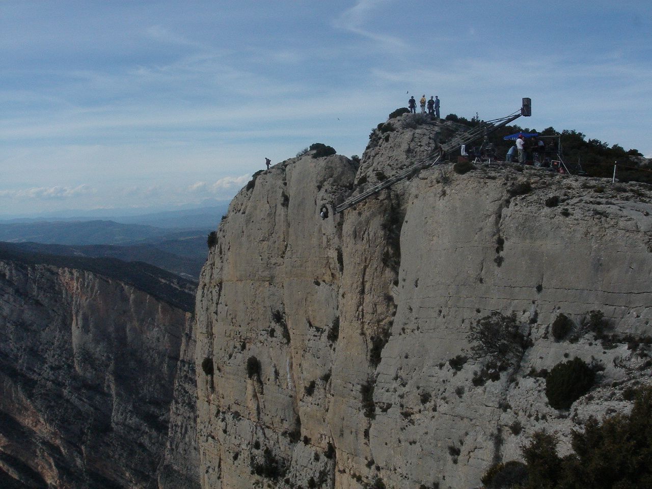 Acantilado rocoso con gente en la cima, cielo azul y colinas distantes al fondo.