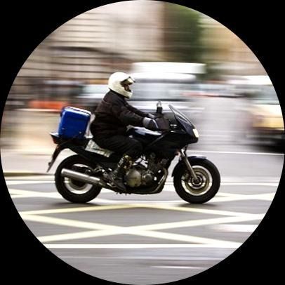 Motociclista con casco blanco, circulando por una calle con una caja de reparto en la parte trasera.