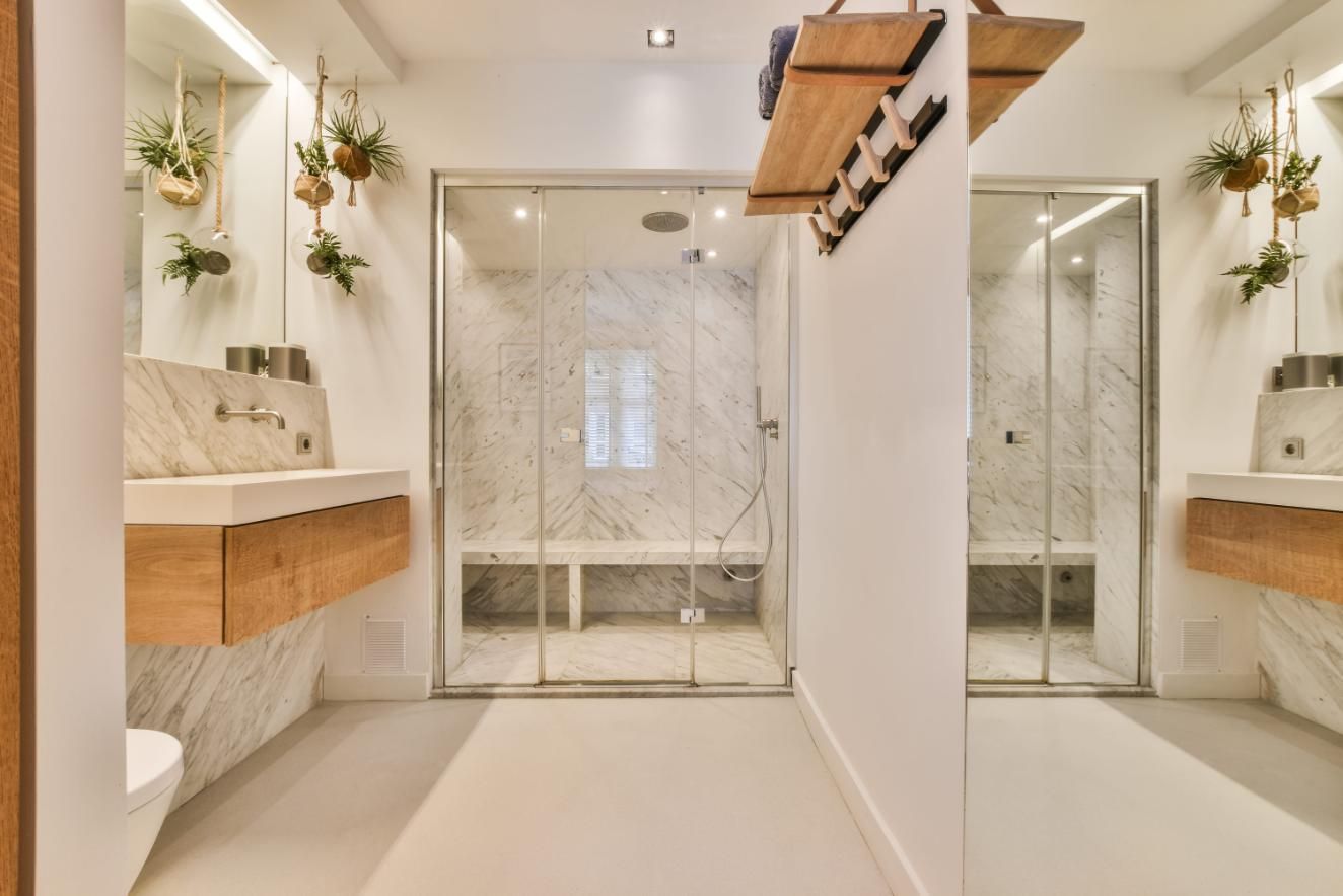 Baño moderno y luminoso con dos lavabos, plantas colgantes y una ducha con mampara de cristal y azulejos de mármol.