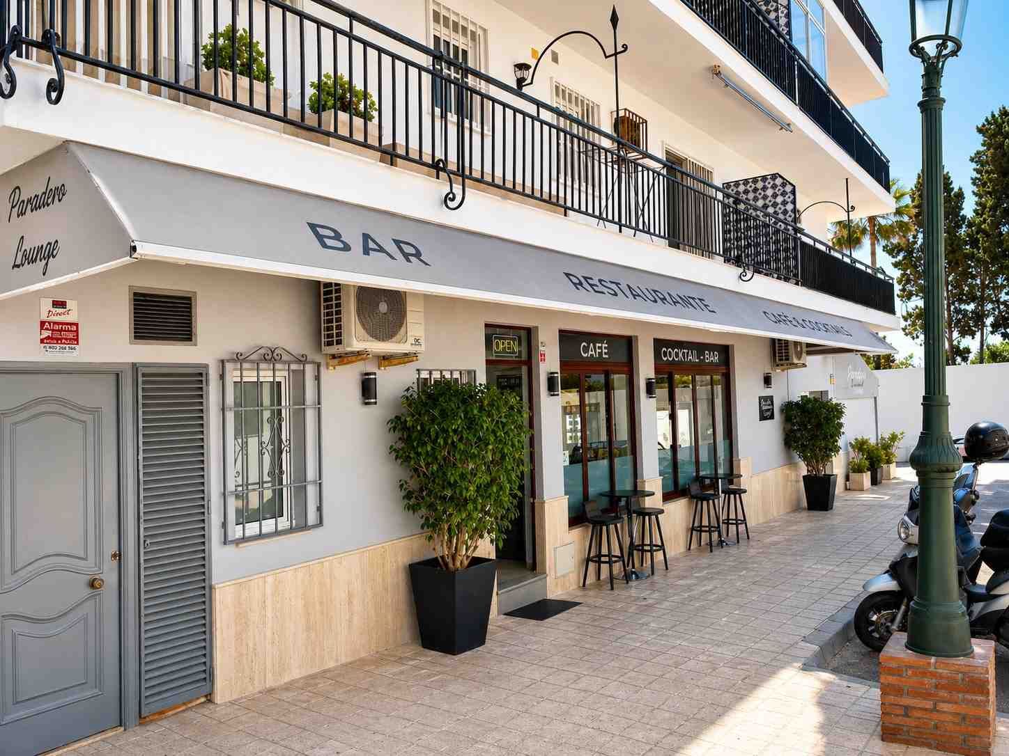 Bar con fachada blanca, toldo a rayas, mesas al aire libre y plantas en macetas en una soleada esquina.