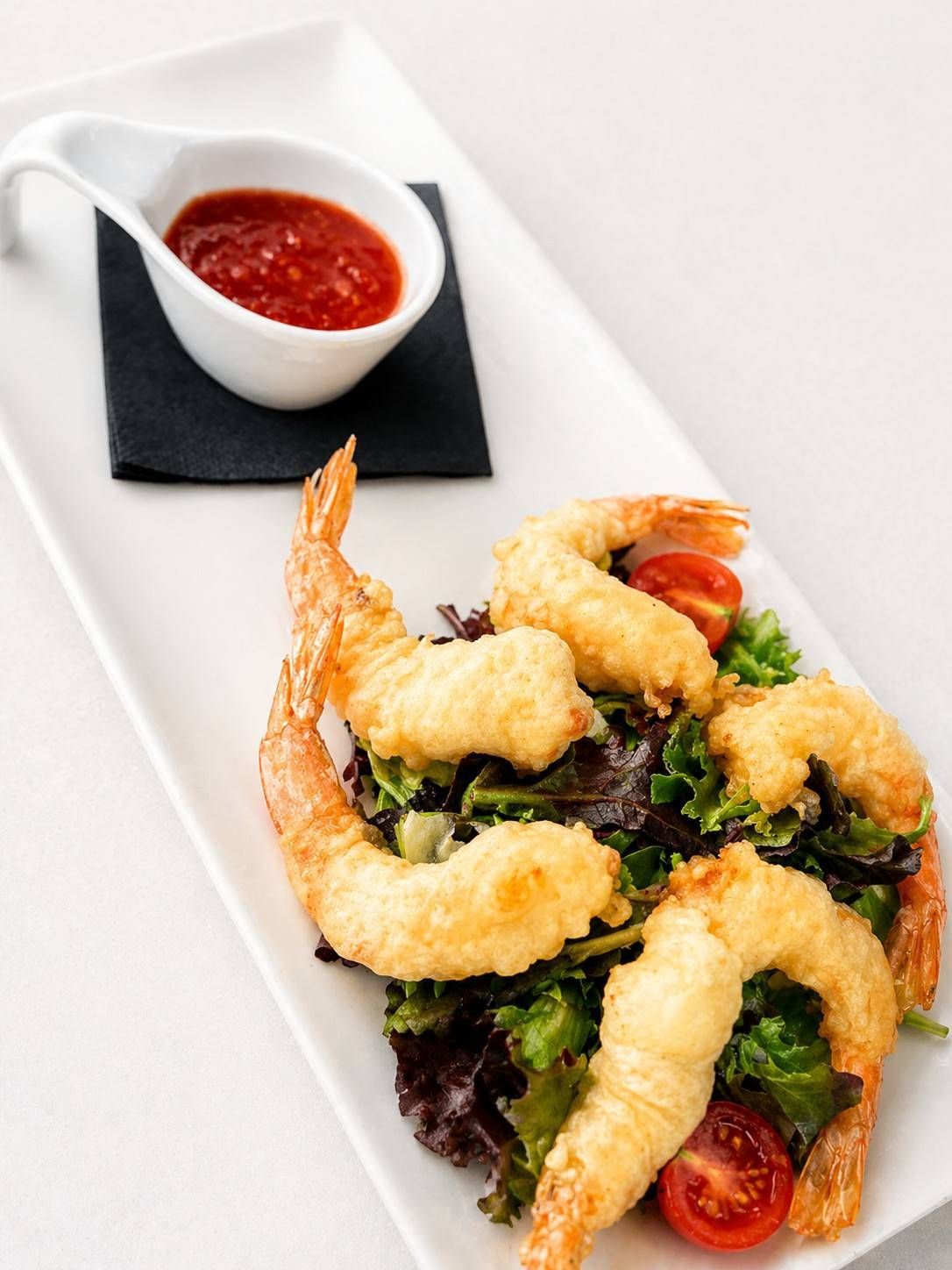 Ensalada de camarones tempura emplatada con tomates cherry y verduras, servida con salsa roja para mojar.