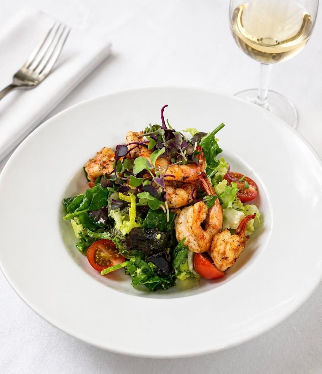 Ensalada con gambas y tomates en un plato blanco junto a una copa de vino blanco.
