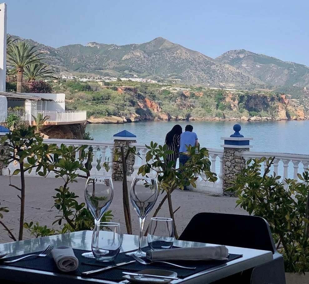 Mesa de restaurante junto al mar con vasos y plantas, con vistas a las tranquilas aguas y las montañas al atardecer.