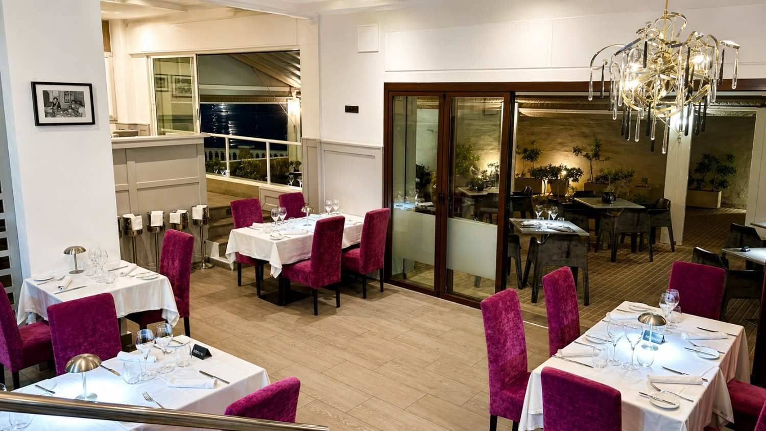 Elegante comedor de restaurante con mesas blancas, sillas magenta y puertas de cristal por la noche.