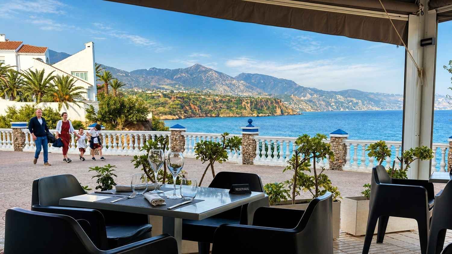 Terraza en un café frente a la playa con vistas al mar azul y a las montañas, mientras la gente pasea por la orilla.