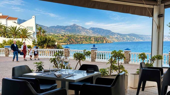 Terraza en un café frente a la playa con vistas al mar azul y a las montañas, mientras la gente pasea por la orilla.
