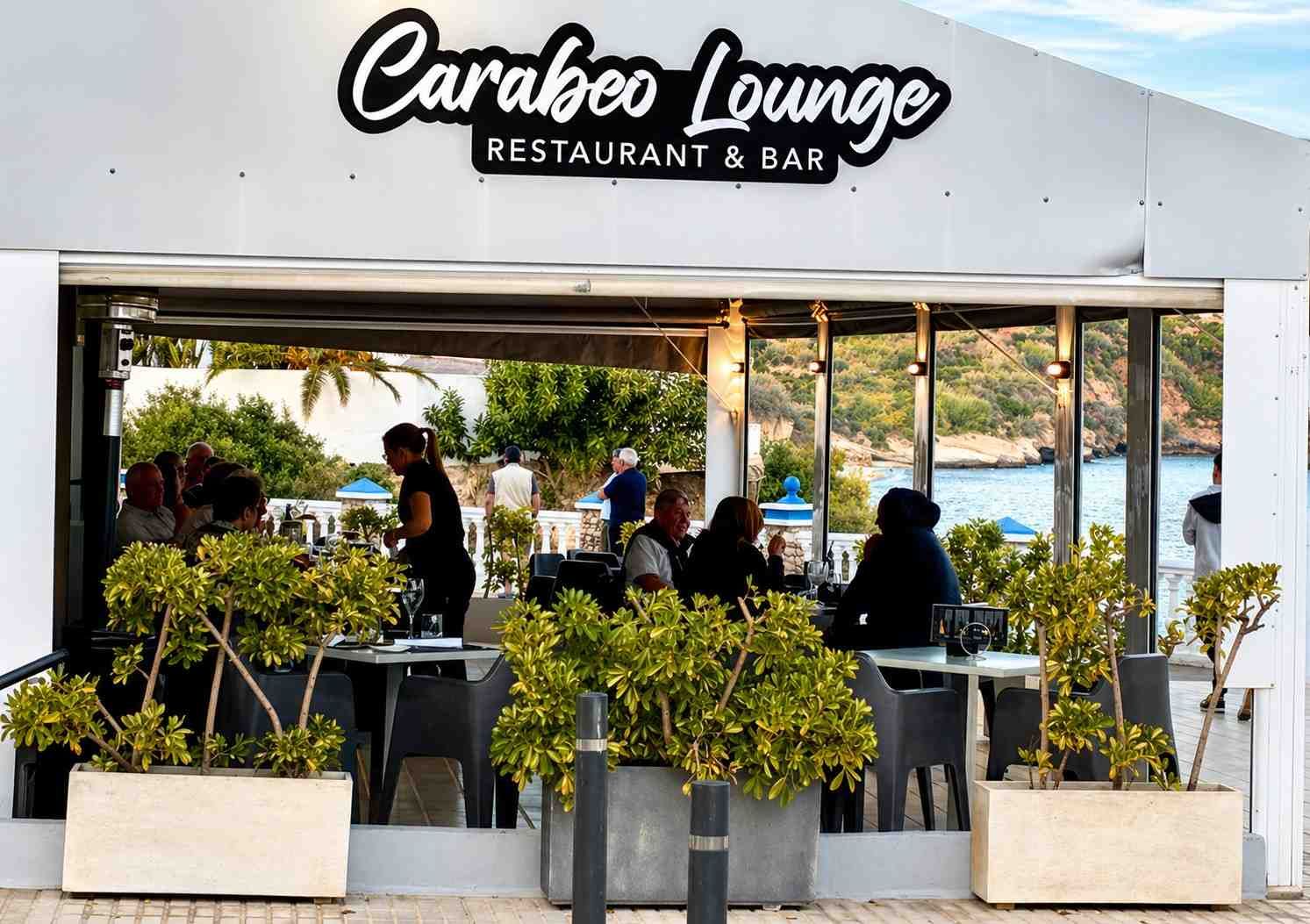 Terraza del restaurante y bar Carabeo Lounge con plantas, mesas y gente con vistas al agua.