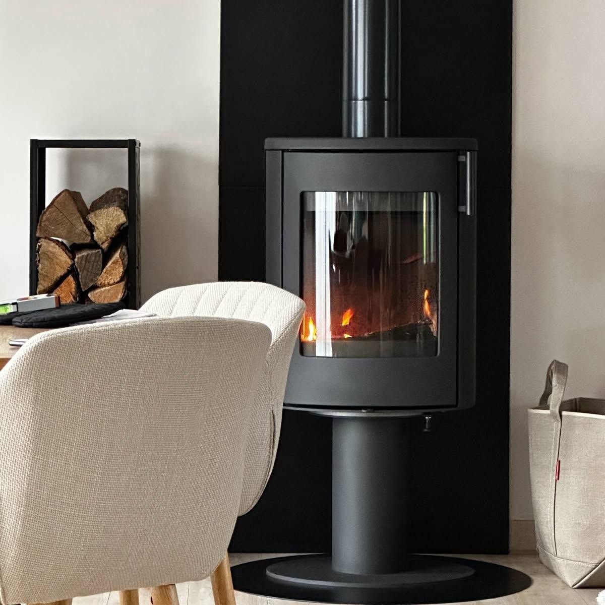 Poêle à bois noir moderne avec feu, à côté d'un fauteuil en osier et de bûches empilées dans une pièce minimaliste