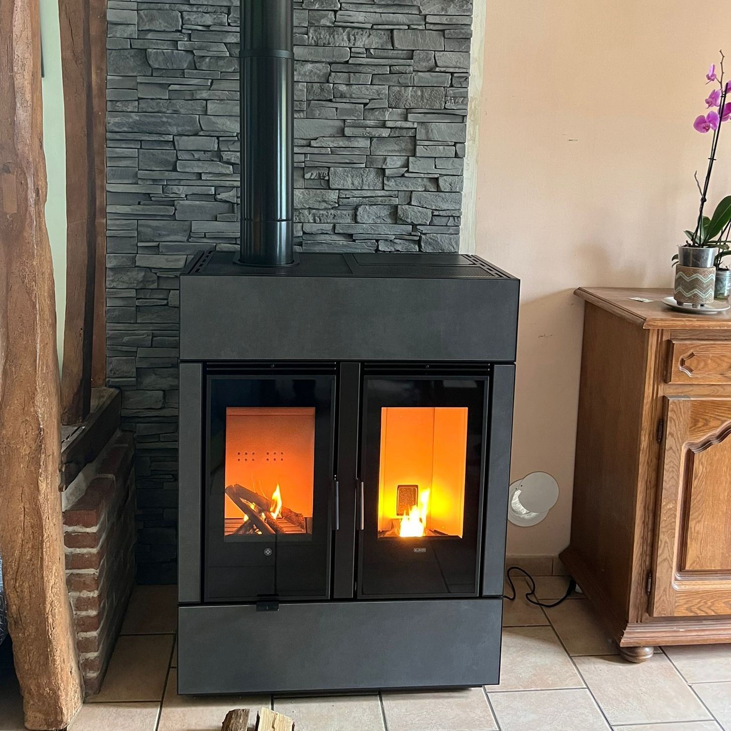 Poêle à bois noir aux flammes visibles dans une pièce confortable, à côté d'une armoire en bois et d'un mur en pierre.