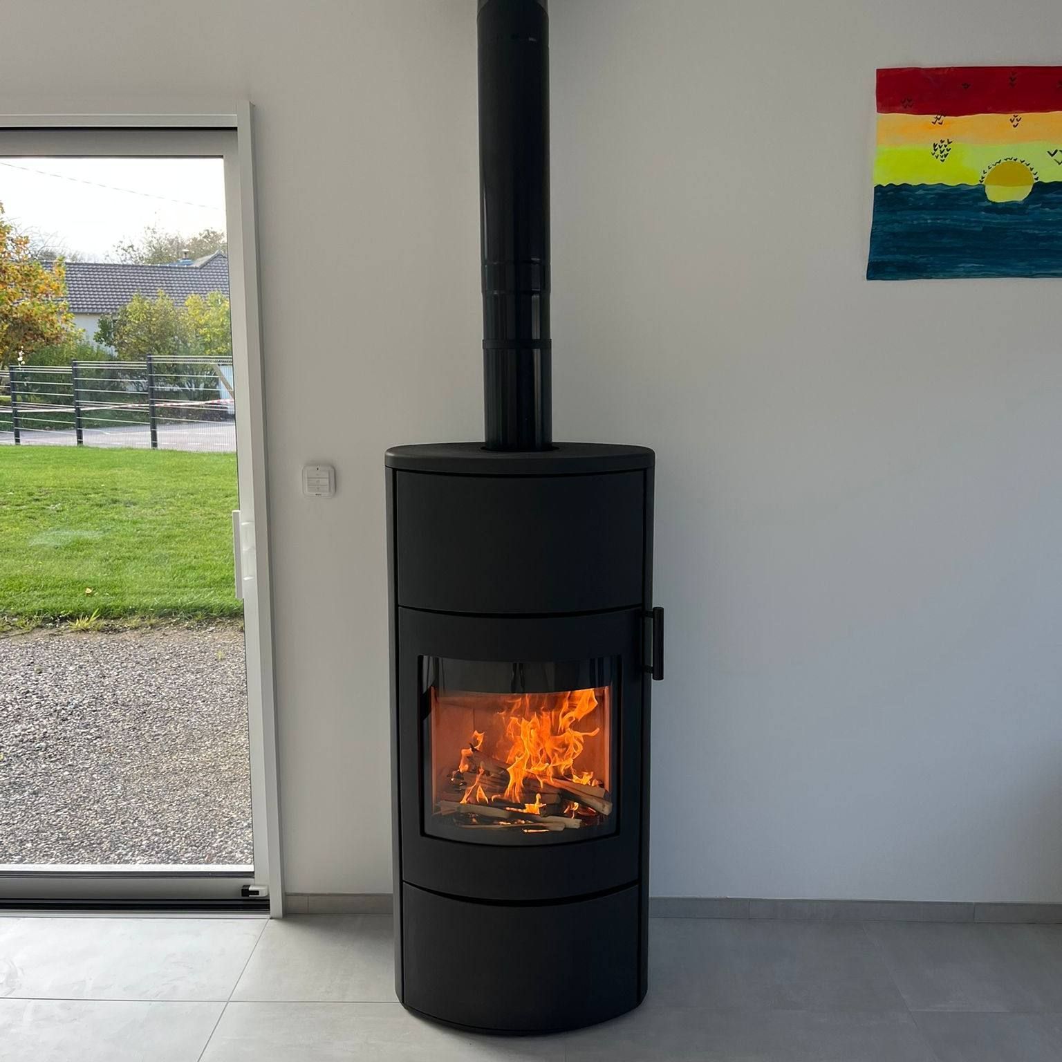 Poêle à bois noir brûlant dans une pièce minimaliste à côté d'une porte vitrée et d'œuvres d'art murales colorées