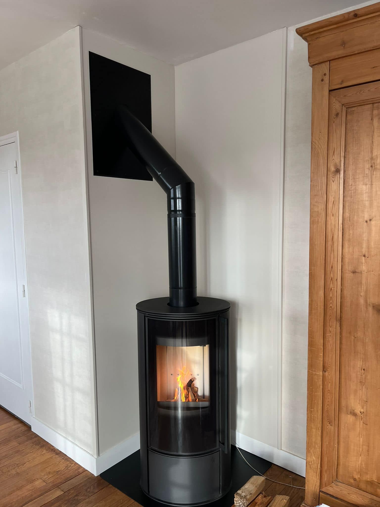 Poêle à bois noir sur pied, flamme visible et conduit de fumée noir, dans une pièce blanche à côté d'un meuble en bois.