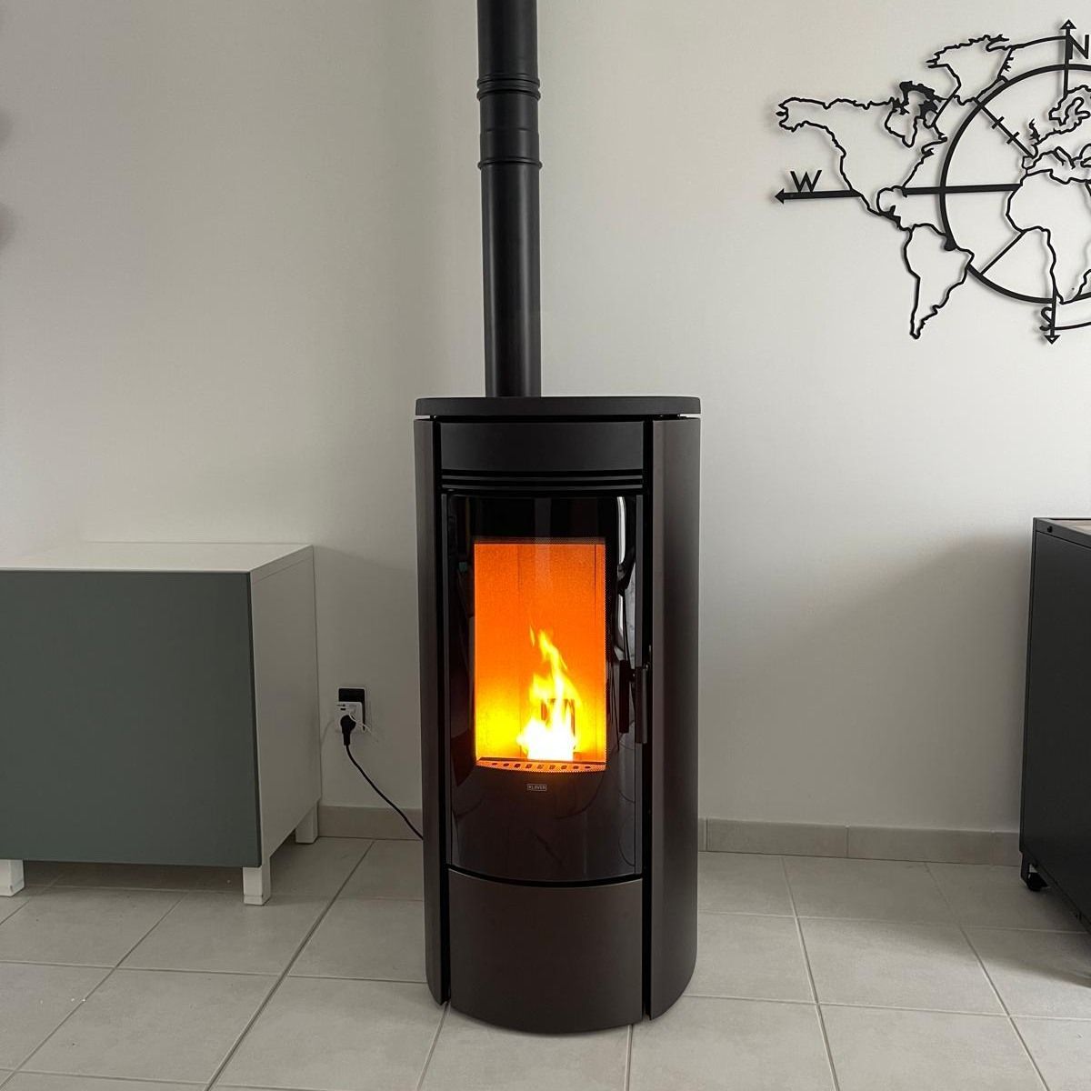 Un poêle à bois noir allumé, avec un feu crépitant, dans une pièce moderne carrelée ornée d'une carte murale.