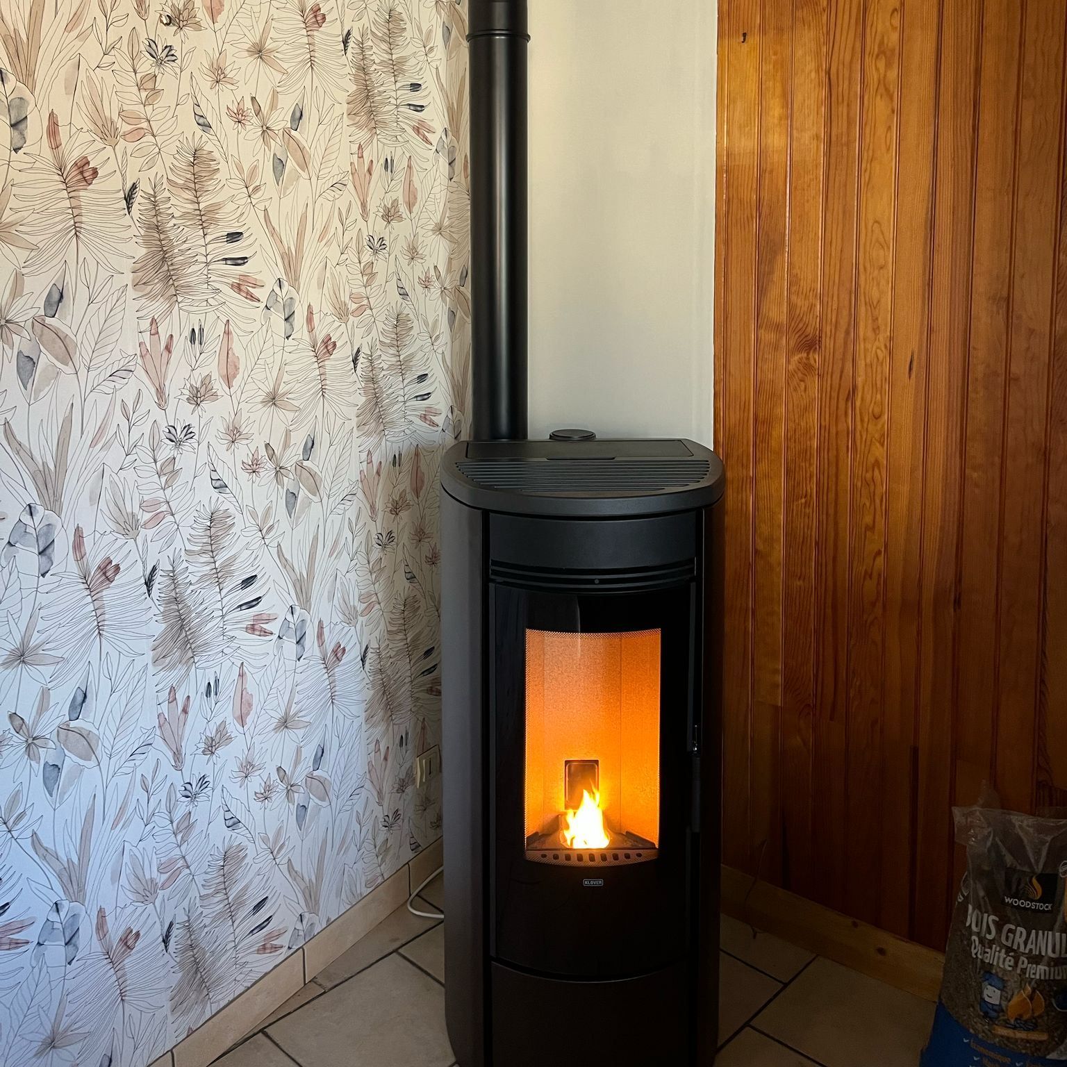 Poêle à bois noir avec un feu orange qui brûle dans un coin, à côté d'un papier peint à motifs floraux et de lambris.