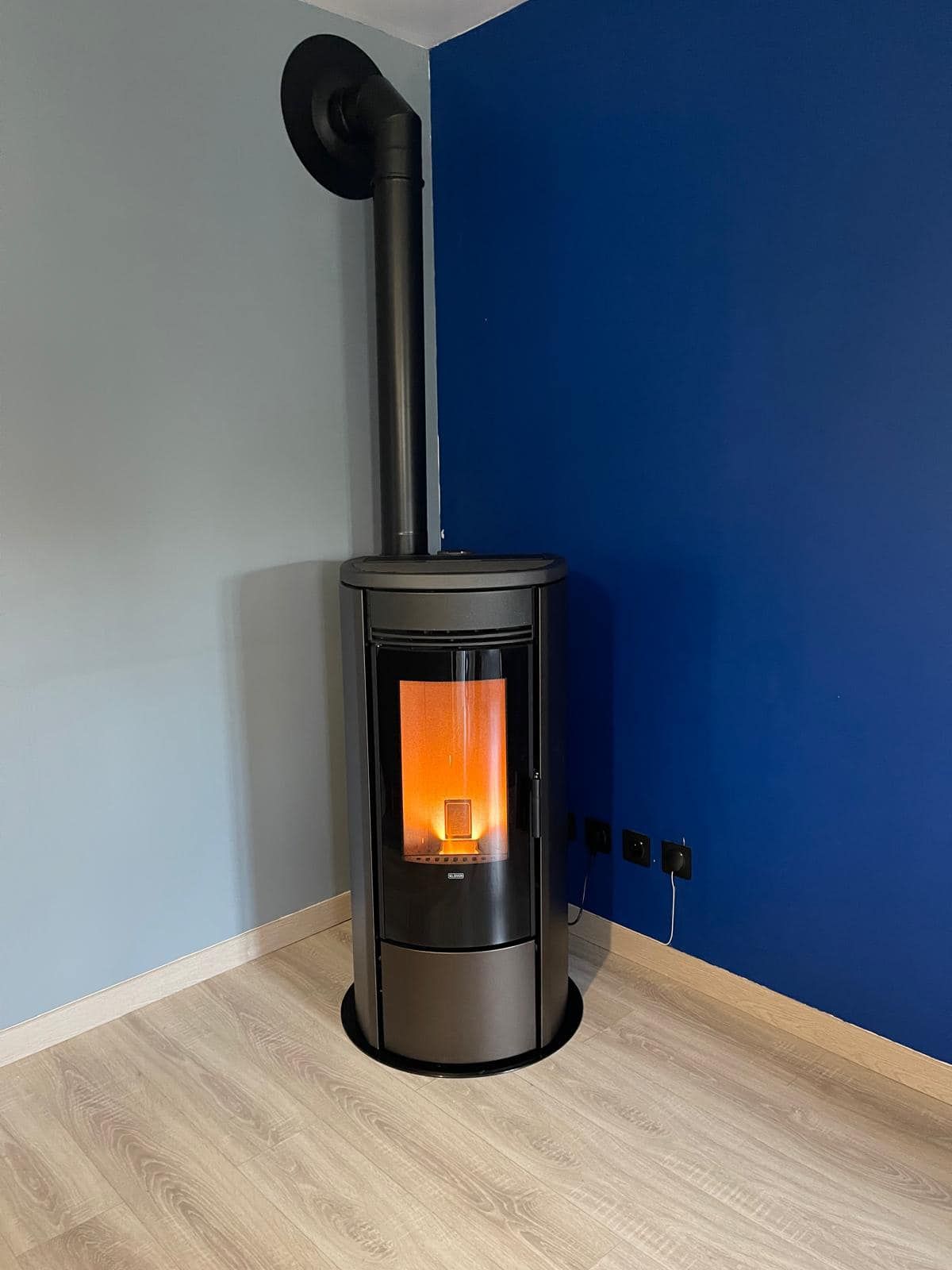 Poêle à granulés noir moderne avec flamme orange dans une pièce aux murs bleus et blancs.