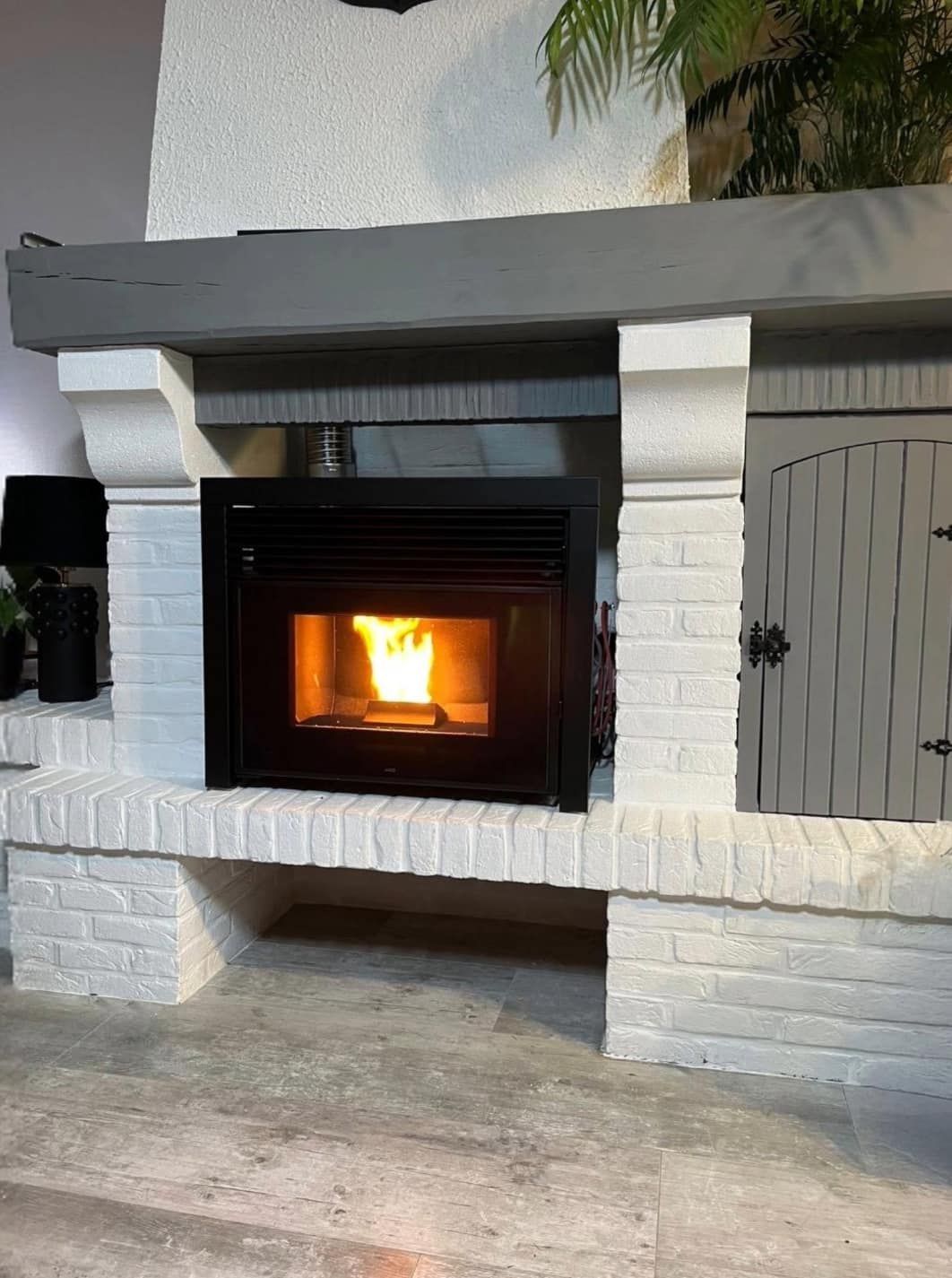 Cheminée extérieure en briques blanches avec un feu allumé et un manteau en bois foncé sous une terrasse couverte