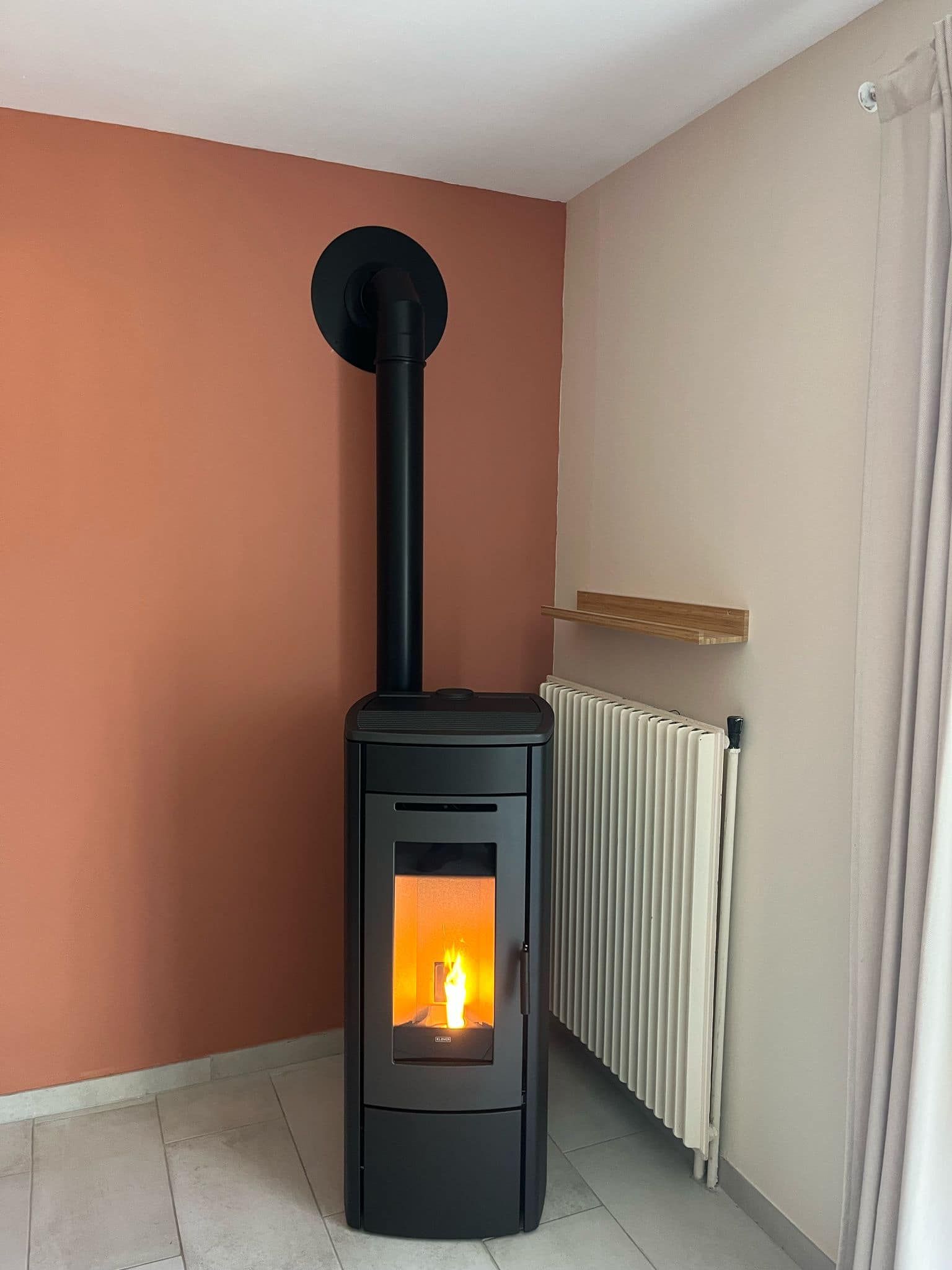 Poêle à bois noir avec un feu allumé dans une pièce d'angle, à côté d'un radiateur et d'un mur couleur pêche.