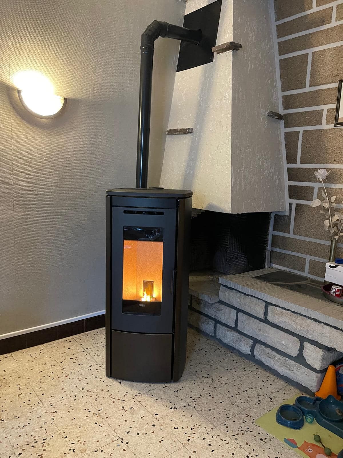 Poêle à bois noir avec un feu crépitant dans un salon carrelé, à côté d'une cheminée en briques et d'une applique murale.