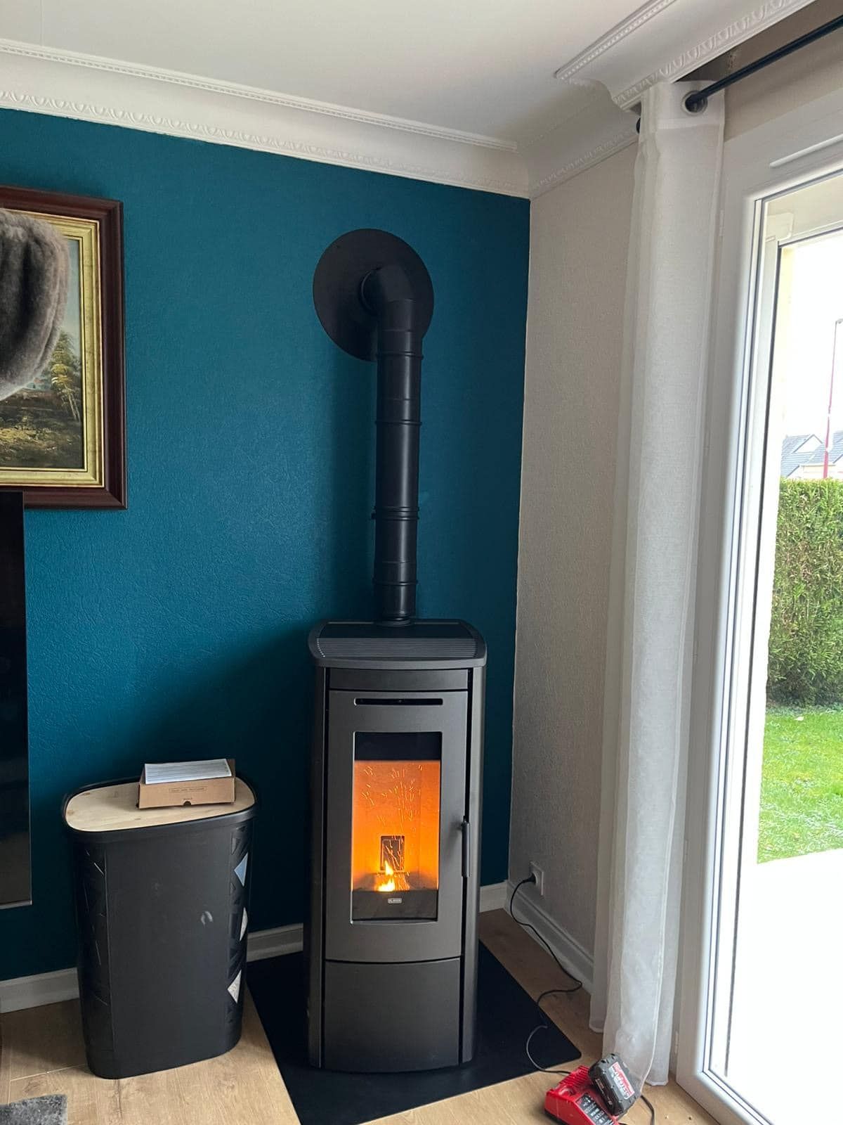 Poêle à bois noir avec flammes orange dans une pièce aux murs turquoise, à côté d'une porte vitrée.