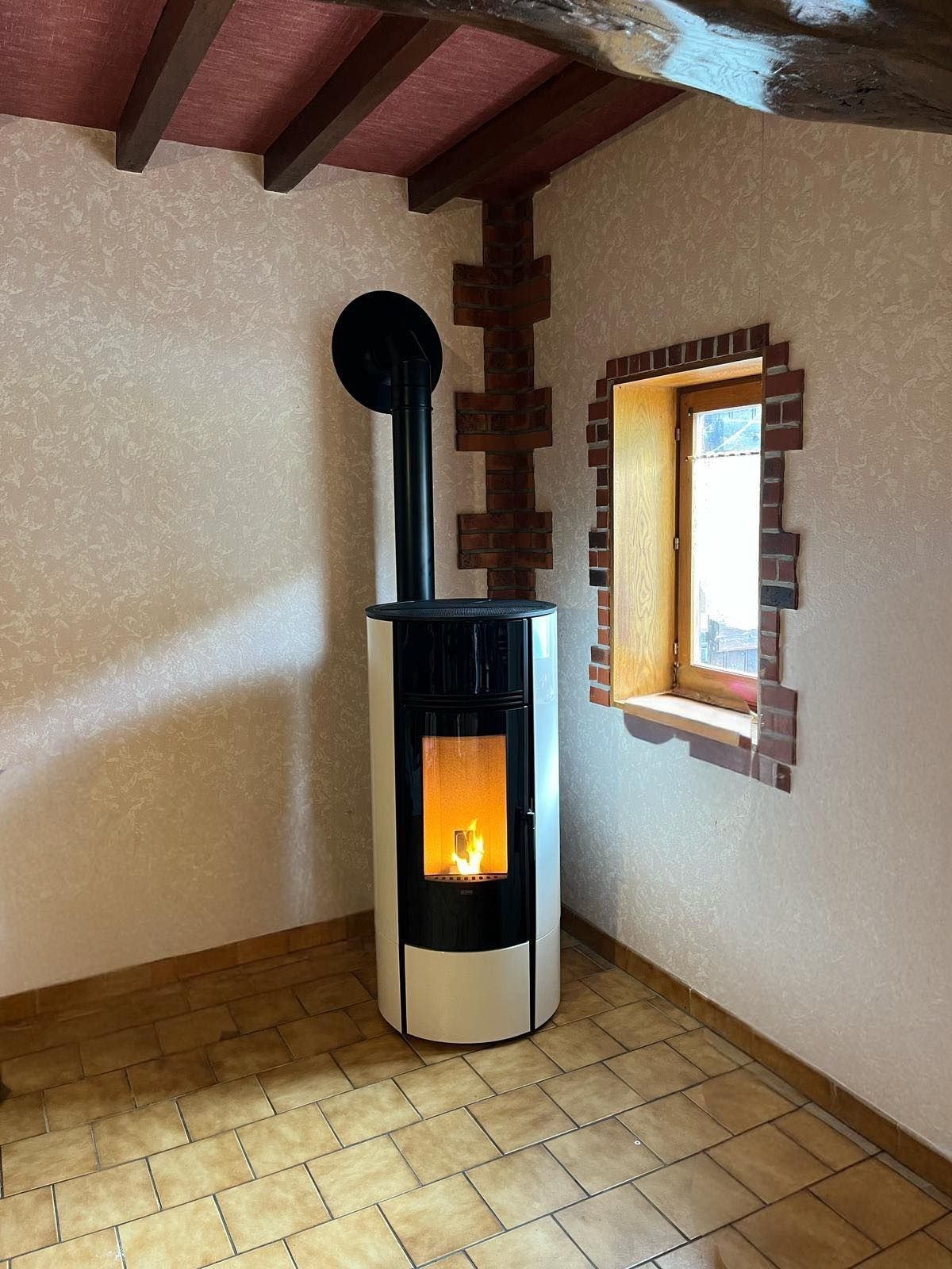 Petite pièce avec un poêle à granulés noirs allumé, un sol carrelé et une fenêtre à encadrement en bois.