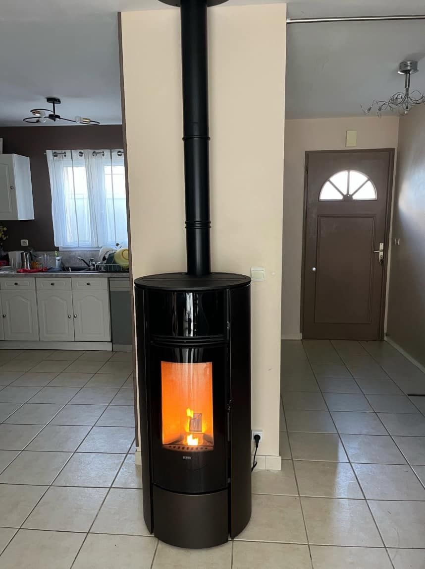 Poêle à bois noir sur pied avec une flamme visible dans un intérieur carrelé, près d'une porte et de la cuisine.