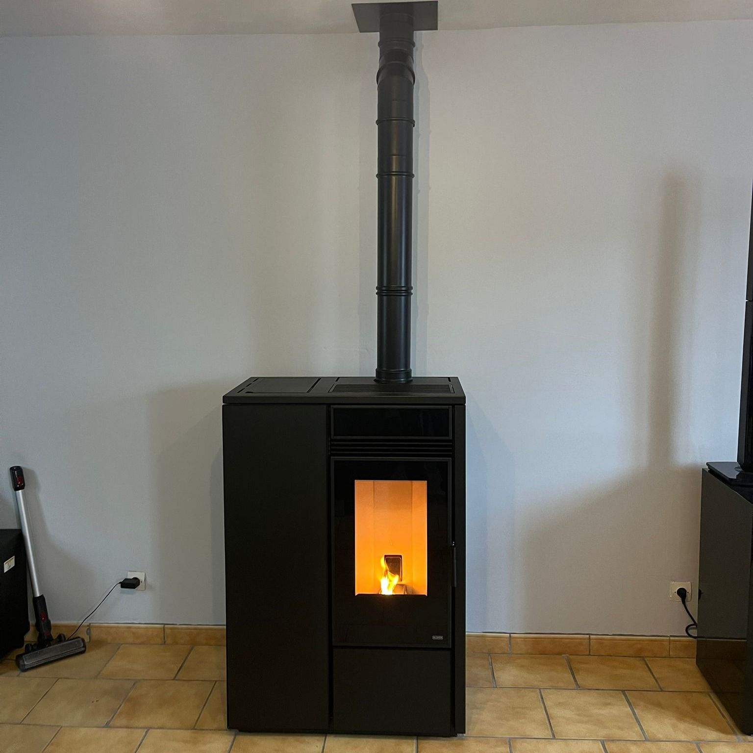 Poêle à bois noir sur pied avec flamme orange, centré contre un mur blanc sur un sol carrelé.