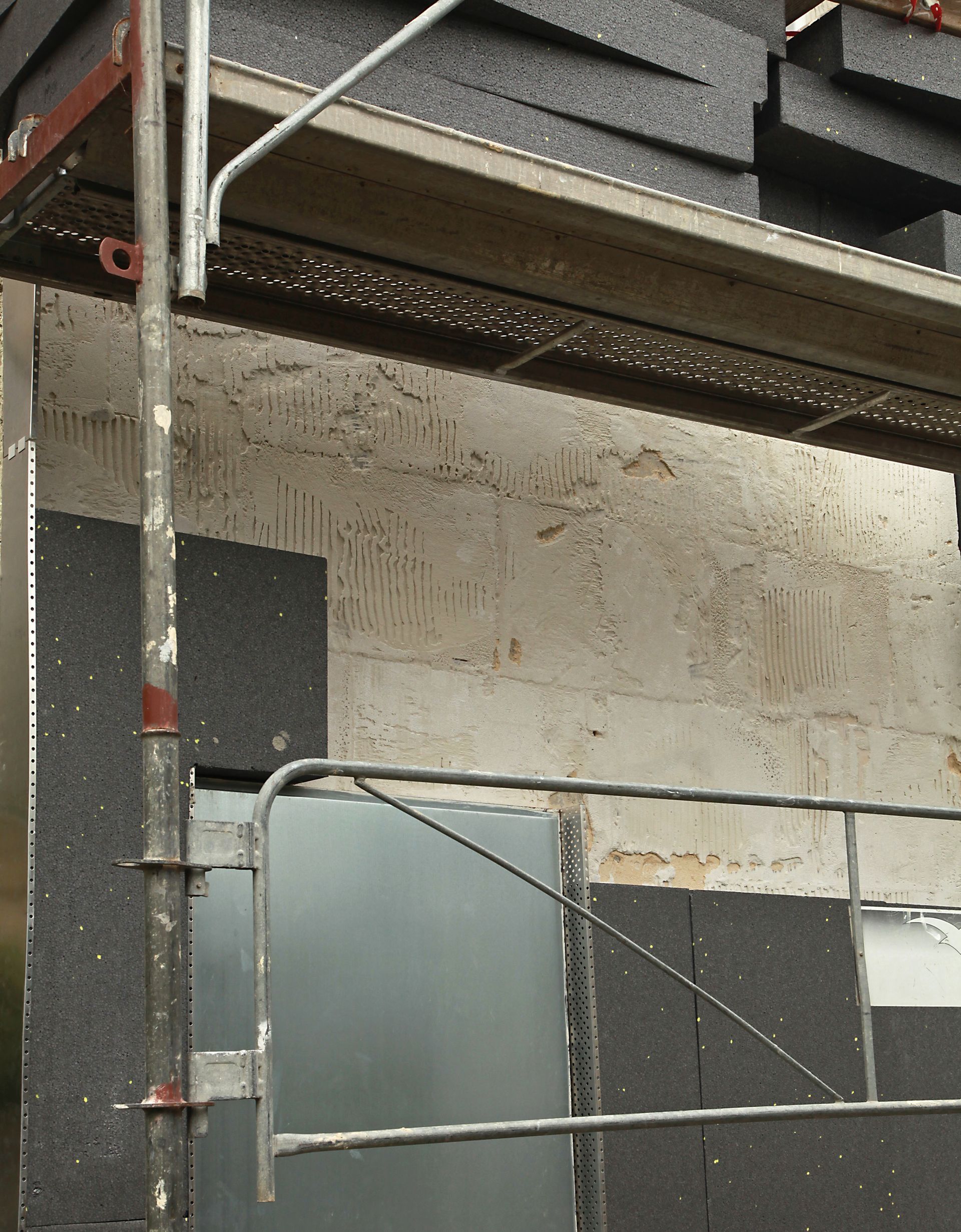 Un chantier montrant des échafaudages positionnés devant le mur d'un bâtiment recouvert de panneaux isolants noirs.