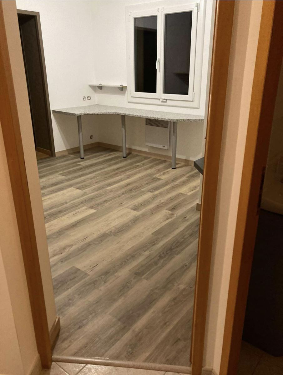 Une pièce avec un sol stratifié imitation bois, des murs blancs et un bureau d'angle placé sous une fenêtre.