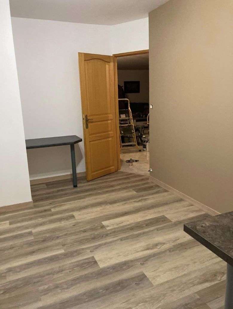 Une pièce avec un sol imitation bois, un mur blanc, un mur taupe donnant sur un couloir faiblement éclairé.
