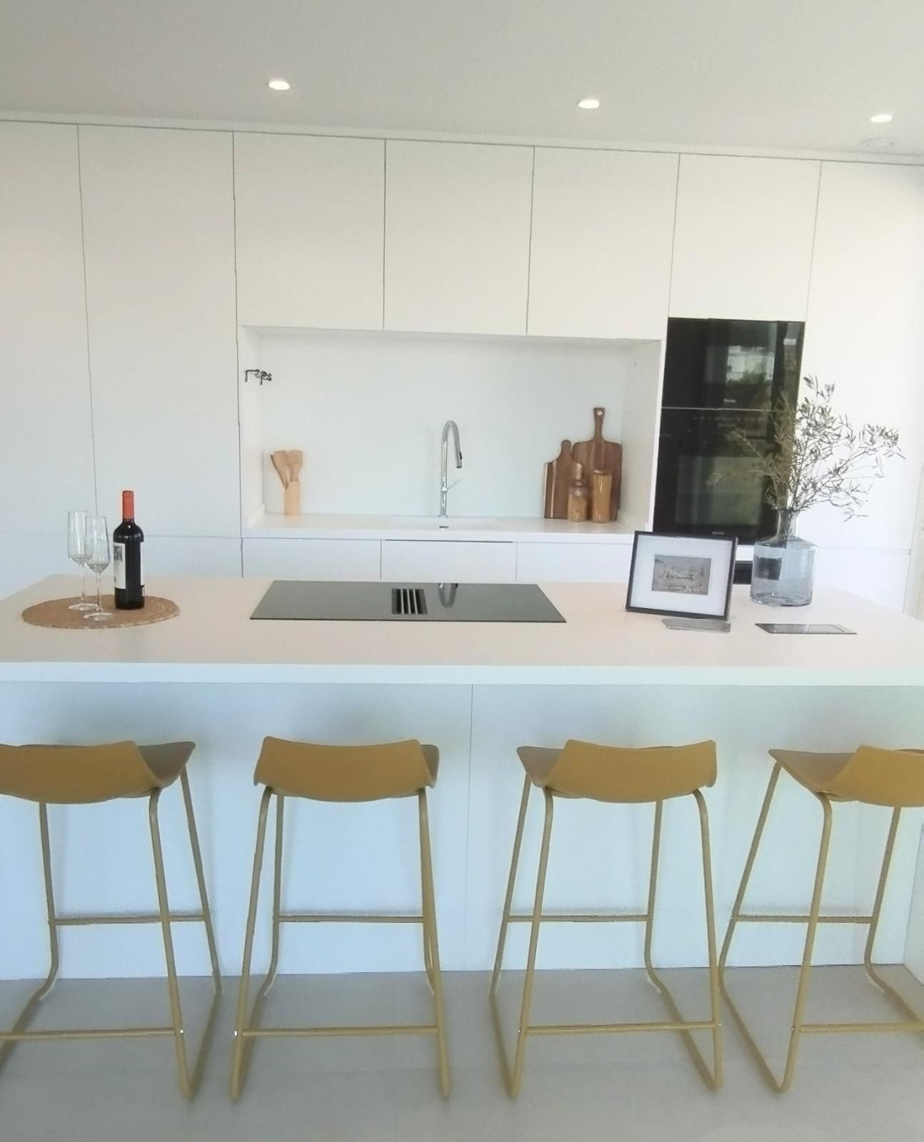 Cocina moderna y luminosa con isla blanca, taburetes dorados, placa de cocina negra y decoración enmarcada sobre la encimera.