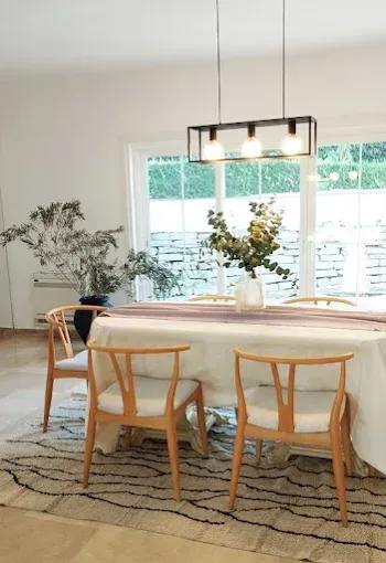 Comedor luminoso con sillas de madera, mesa redonda, lámpara colgante y flores junto a una gran ventana.