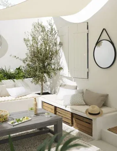 Patio moderno y luminoso con bancos blancos, árbol en maceta, mesa gris y espejo de pared redondo.