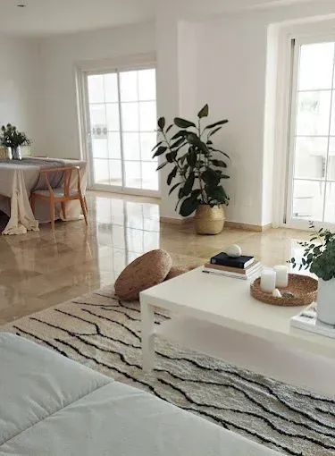 Luminoso salón moderno con mesa de centro blanca, alfombra, planta de interior y zona de comedor junto a grandes ventanales.