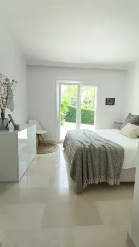 Habitación luminosa y minimalista con cama blanca, suelo de baldosas claras y puertas francesas que dan a un jardín verde.