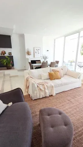 Sala de estar luminosa y moderna con sofá blanco, sillas grises, chimenea y grandes ventanales.