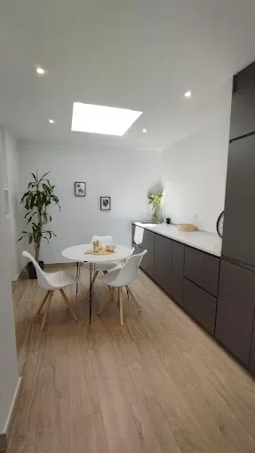 Comedor moderno y luminoso con mesa redonda, sillas blancas, suelo de madera y armarios oscuros a lo largo de la pared.