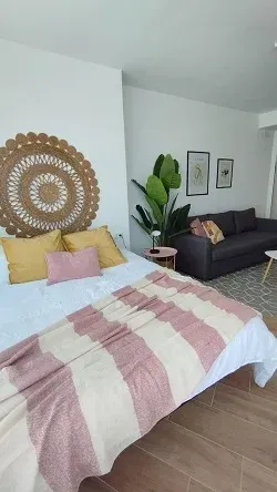 Habitación luminosa con colcha a rayas, cuadro de pared tejido, plantas de interior y un sofá oscuro junto a la cama.