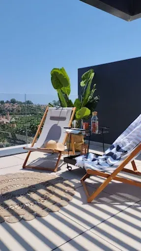 Terraza soleada en la azotea con tumbonas de madera, plantas en macetas y vistas a la ciudad tras una pared negra.