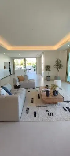 Sala de estar moderna y luminosa con sofá blanco, alfombra, mesa de centro y grandes ventanales que dan a un patio soleado.