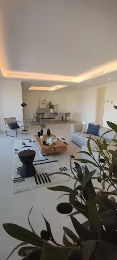 Sala de estar moderna con sofás blancos, plantas, alfombra en blanco y negro y luces empotradas cálidas en el techo.