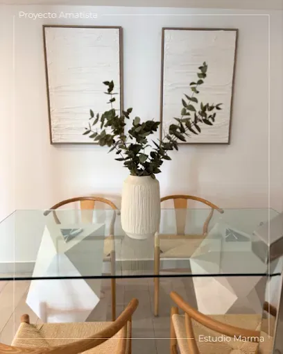 Mesa de comedor de cristal con sillas de mimbre y un jarrón blanco con plantas debajo de dos cuadros enmarcados.