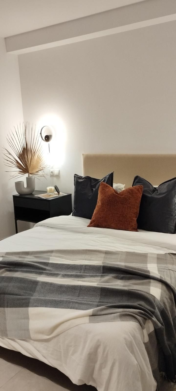 Habitación acogedora con cama hecha, manta de cuadros, almohadas oscuras, lámpara de noche y decoración neutra.