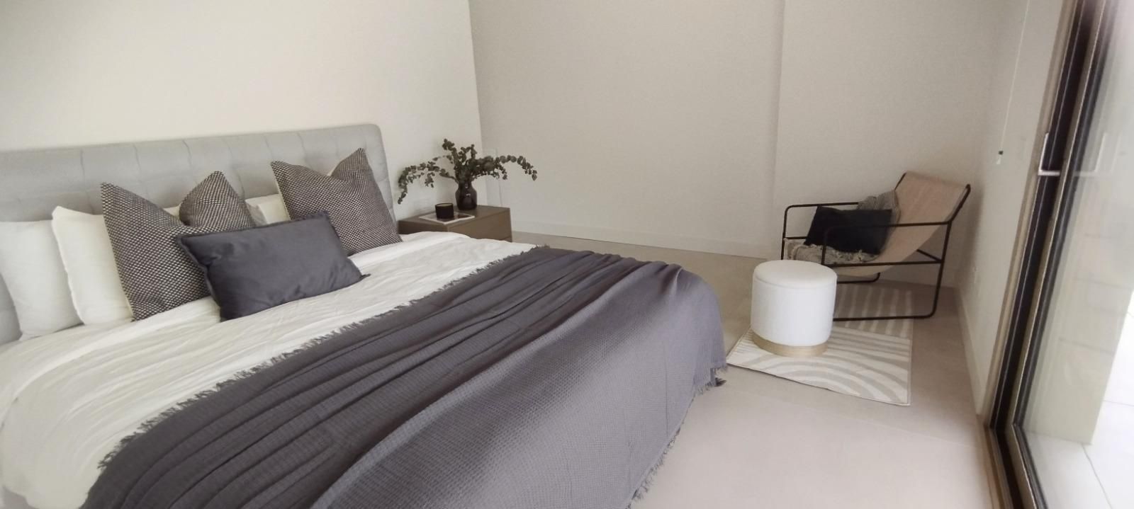 Dormitorio moderno y luminoso con una cama gris bien hecha, almohadas, silla auxiliar y puertas corredizas de cristal.